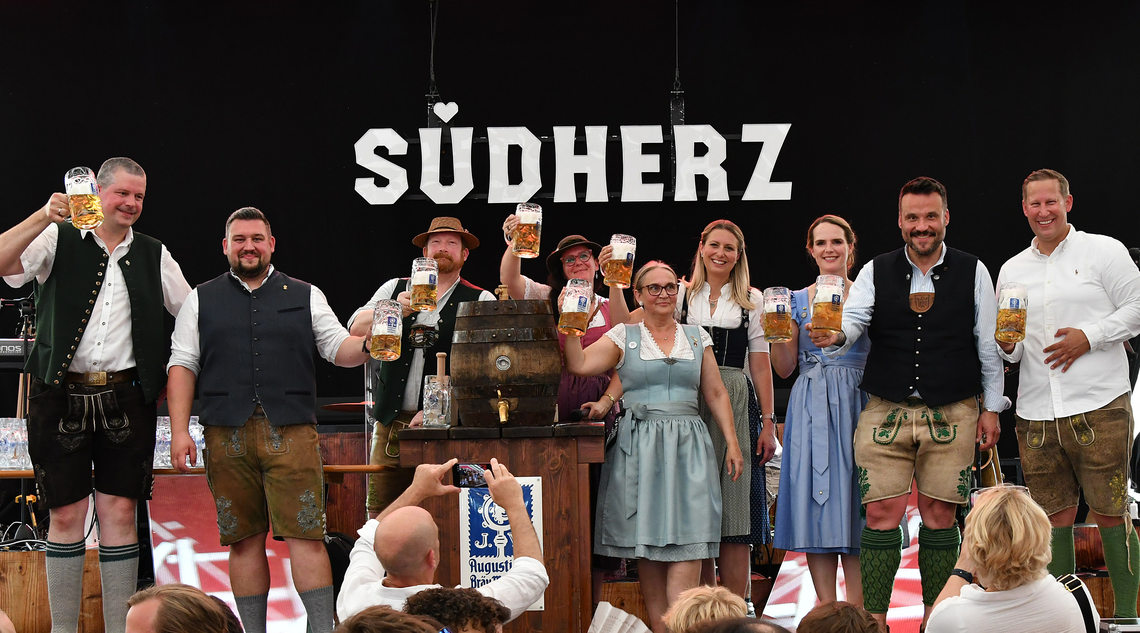 Mehrere Personen stehen mit erhobenen Gläsern auf der Bühne und prosten der Menge im Publikum zu.