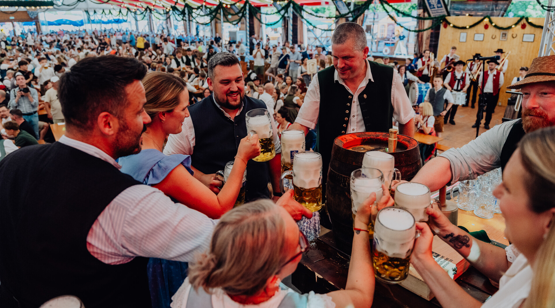 Mehrere Personen prosten sich zu. Im Hintergrund sitzen die Festzeltbesuchenden auf Bierbänken.