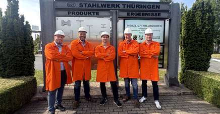 Fünf Personen mit orangenem Kittel und weißen Helmen vor einer Tafel mit der Beschriftung "Stahlwerk Thüringen"