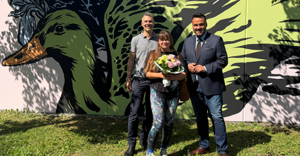 Zwei Männer und eine Frau mit Blumenstrauß stehen vor einer Wand, die mit einem Vogel bemalt wurde.