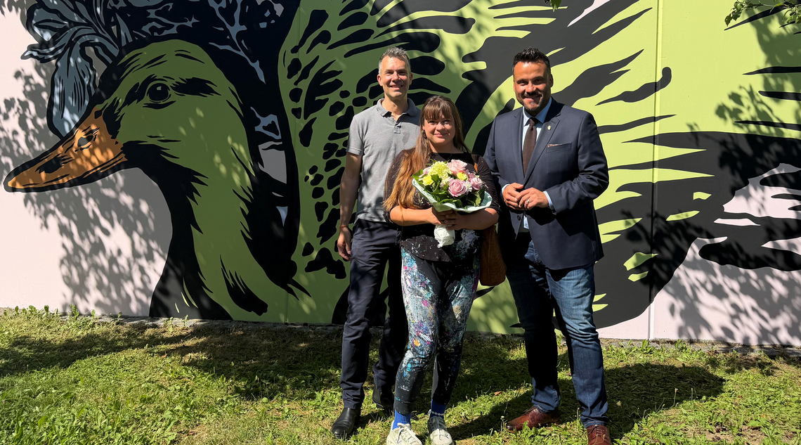 Zwei Männer und eine Frau mit Blumenstrauß stehen vor einer Wand, die mit einem Vogel bemalt wurde.
